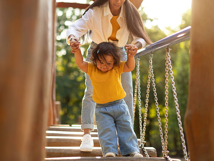 Mother Holding Daughter's Hands Walking Swinging Bridge On Playground