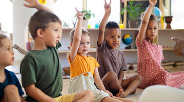 group of young children sitting on floor indoors in classroom raising hands
