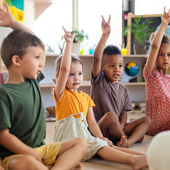 group of young children sitting on floor indoors in classroom raising hands