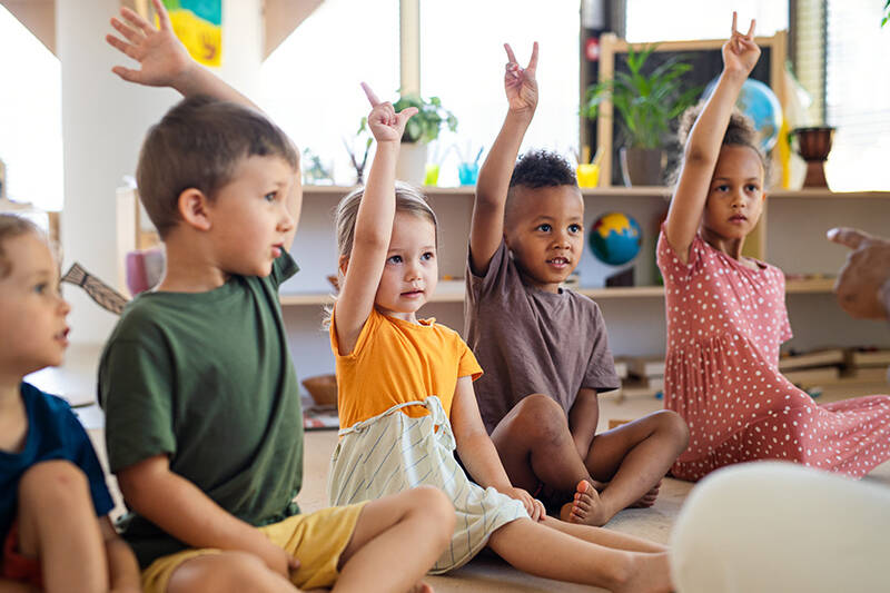group of young children sitting on floor indoors in classroom raising hands